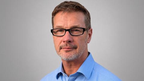 Close-up man with greying hair and greying beard, wearing glasses and a blue shirt