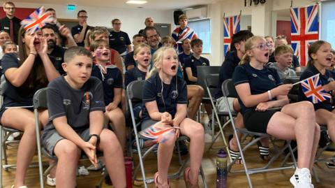 Boys, girls, women and men wearing blue T-shirts, holding Union flags, sitting on chairs, and cheering Maisie Summers-Newton on as they watch her take part in the Paris Paralympics 2024