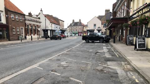 Pocklington Market Place. The road surface in the parking bays is marked with potholes. There is only one vehicle parked in the parking bays. The Black Bull pub can be seen. 