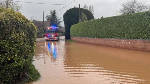 A red fire engine with its blue lights on. it is attempting to cross a brown body of flood water. there are green hedges on either side of the flood water