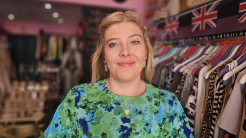 Tilly wears a green and blue floral dress and is smiling at the camera. She had a heart shaped silver necklace on and light blue dangly ear rings. Her hair is blonde and shoulder length, it's down but clipped off her face. Behind her is a rack of clothing and union jack bunting.