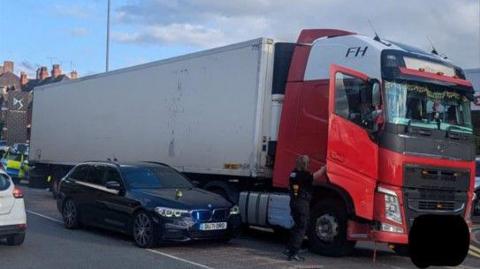 A lorry stopped on the road with a car next to it and the driver's door open, as a woman in black gestures up