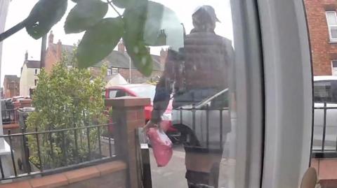 Doorbell camera footage of a man dressed in black with a black cap walking out of a front garden with a pink parcel.