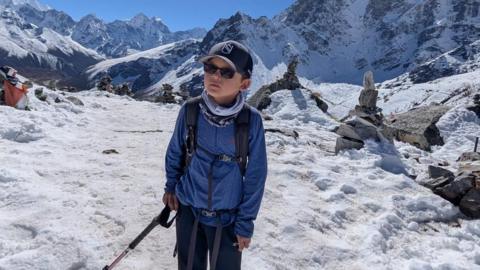 Rio stood in the Himalayan mountains. He is wearing a navy cap, holding a walking stick, and has a blue coat on.