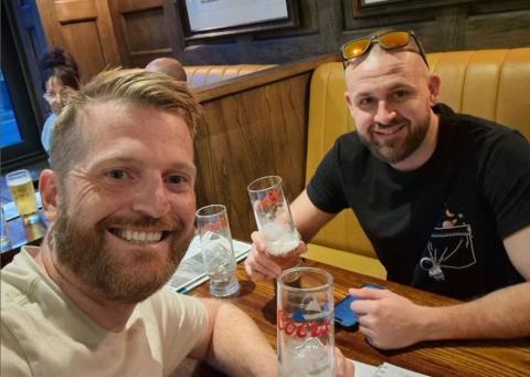 James Askew, in a beige T-shirt, holding a glass and sitting opposite Will Shreeve, who is holding also  a glass and wearing a black T-shirt.