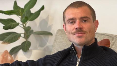Ross is sitting on a light-colored couch in a modern living room, wearing a dark zip-up sweater. A green potted plant is visible behind the couch, along with decorative pillows and a framed picture partially in view. The setting appears calm and contemporary.
