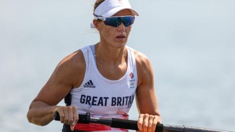 Helen is wearing a white cap with a Team GB logo and a white Team GB vest holding an ore