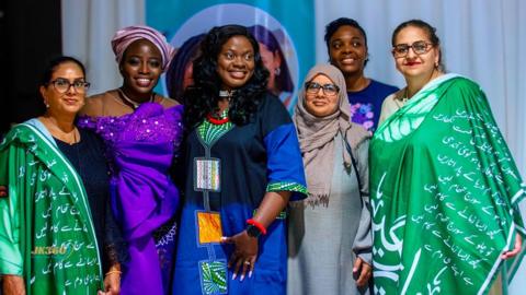 A group of black and South Asian ladies, all dressed in colourful costumes which represent their different cultures. The black ladies wear African prints, some of the South Asian ladies wear shawls with Urdu writing on them