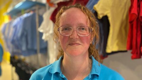 Kim Ansell is wearing glasses and a blue shirt with racks of school uniform hanging neatly behind her