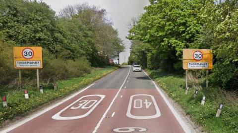 The A19 as it enters Thormanby with 30mph signs each side of the road and speed limit markers in white on the road and a Thormanby village sign post.