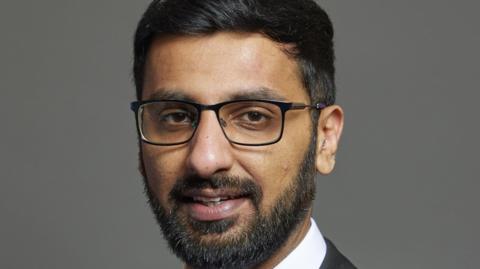 Official portrait of Adnan Hussain taken against a grey backdrop. He has black hair and a beard and is wearing glasses.