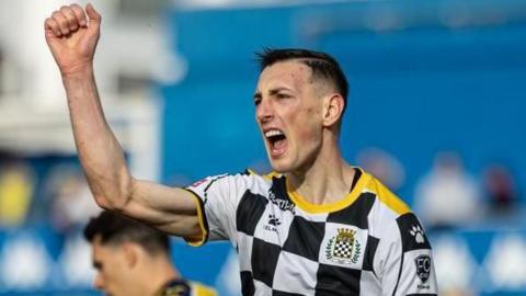Robert Bozenik in a black-and-white checked Boavista shirt celebrates a goal by putting his right fist up above his head