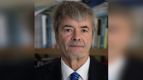 A man, Prof Julian Elliott, with grey hair and a beard wearing a black suit, white shirt and stripy blue and black tie. There are bookshelves in the background.