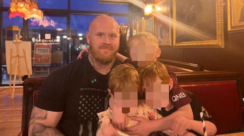 Lenny Scott, who has a bald head, ginger beard and tattoos on his neck and arms, is photographed in a restaurant while holding his three children. Their faces have been obscured in the photograph released by Lancashire Police.