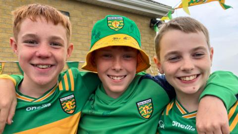 Three young boys in Donegal GAA gear. The boys on the left and right are wearing yellow and green county jerseys while the boy in the middle, who has his arms around the other two, is wearing a green Donegal jumper and green and yellow bucket hat.
