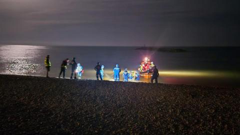 Coastguard and lifeboat crews stand on Sidmouth beach during a rescue operation to bring a small dinghy back to shore during the night. A person is being brought back to the beach by two rescuers who are knee-deep in the water. An orange lifeboat with a light shining is anchored just off the beach.
