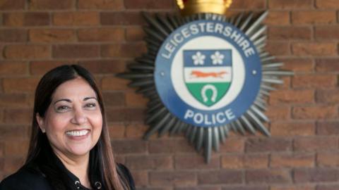 Rani Mahal is smiling at the camera. The Leicestershire Police badge is in the background against a brick wall.