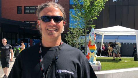 Dr Martin Khechara smiles while holding a booklet about the festival in front of a gazebo and inflatable balloon entrance way. He is wearing a black T-shirt and sunglasses