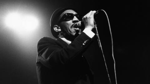 A black and white image of Laurel Aitken wearing a hat and sunglasses, singing into a microphone