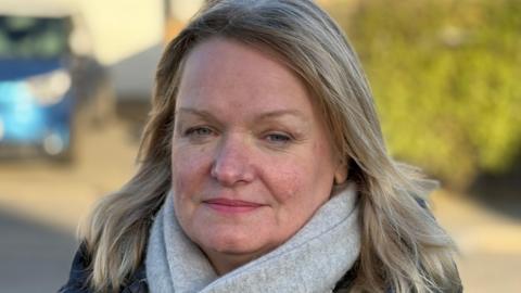 A woman with shoulder length blond hair with a light grey coloured scarf around her neck in a dark coat. 