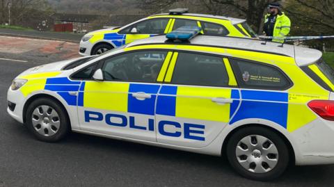 Two police cars are parked parallel to each other on a road. A uniformed officer stands at the back of one next to police scene tape. Some houses can be seen in the background.