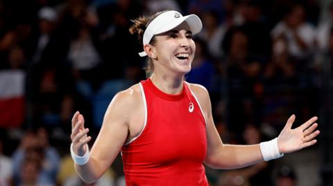 Belinda Bencic, in red tennis top and white visor, celebrates with a smile and arms spread wide, with crowd visible behind her