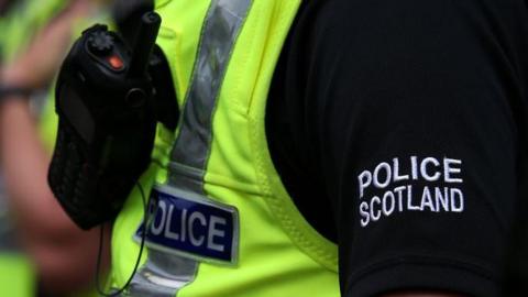 Close up of Police Scotland officer in uniform with yellow hi-vis and walkie-talkie type device.