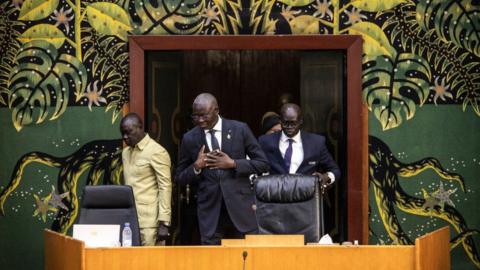 Senegal President Macky Sall wears a black suit and stands with his hands on his chest in the National Assembly, with other ministers walking into the room behind him