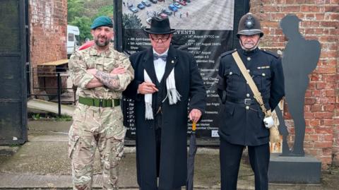 Three men dressed in different outfits - one as a Royal Marine wearing a green beret, one as Winston Churchill, and another in a vintage police officer uniform.