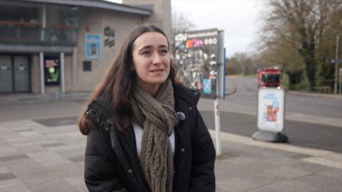 Young womanwith dark hair wearing coat and long scarf standing in front of a concrete building
