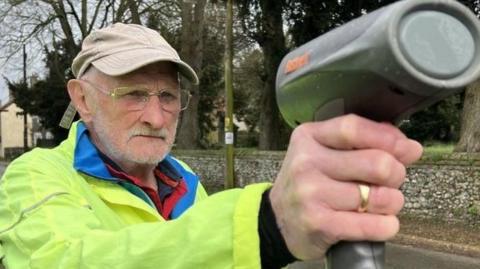 Stock image of Alan Petto, from Great Massingham, holding a speed gun. He wears a high visibility yellow coat, beige cap and glasses. He has grey hair and facial hair and wears a gold ring on the hand holding the speed gun.