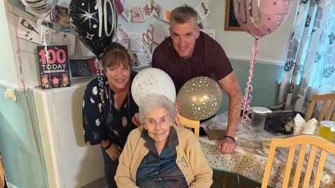 Elderly woman wearing a yellow cardigan sitting with relatives amongst party decorations