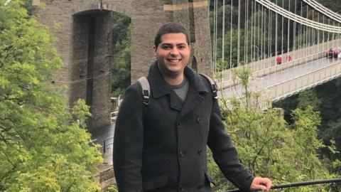 Itamar Bareket, a man with dark hair and a bright smile, he wears a black formal coat and a rucksack, with stands in front of a grand bridge.