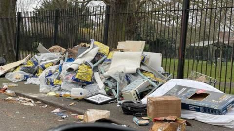 Fly-tipping in Southall on the pavement. Rubbish is piled up to half the height of the tall fence behind it. Items include picture frames, carpets, plastic bags and cardboard.