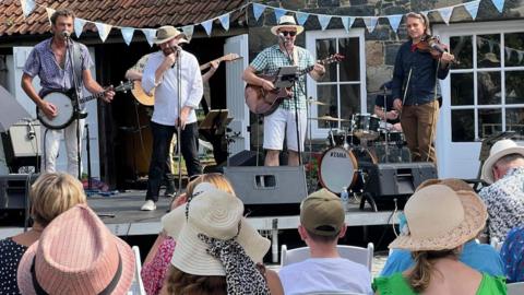 A band plays on a stage in front of rural building, listened to by a crowd on seats.