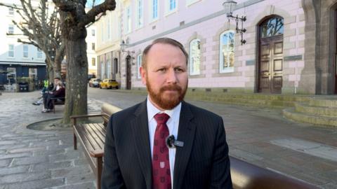 Deputy Tom Coles is wearing a black blazer and a red tie. He is sitting in Jersey's Royal Square. 