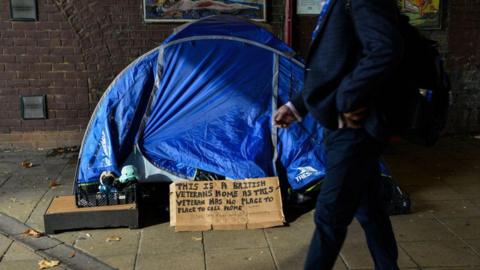 A man walks past a tent on a pavement, which has a cardboard sign outside it reading: "This is a British veterans home as this veteran has no place to call home"