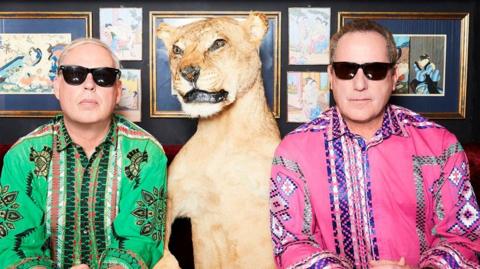 Two men wearing black sunglasses are pulling serious poses whilst wearing very colourful shirts with patterns/ One man has short blond hair and a green shirt and the other on the right has a pink shirt and short brown hair. In the middle is a taxidermy lion and there are photos on the wall behind them.