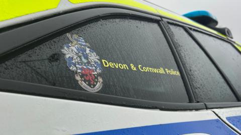 The outside of a Devon & Cornwall Police car with the force's logo printed on the window. It's raining with lots of water running down the window. 