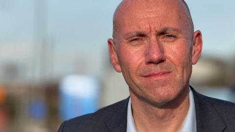 A smartly dressed man with a shaved head wears a serious expression as he looks into the camera. He is wearing a dark suit and light-blue shirt. The background is out of focus and shows a blue sky and blurred buildings.