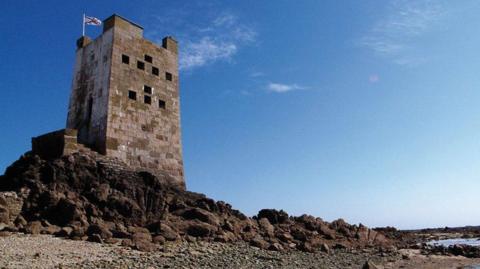 A photo of Seymour tower, surrounded by rocks at low tide. The sky is blue. 