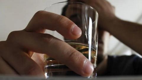 A man holds a drink in his hand close to the camera with his other hand on his head
