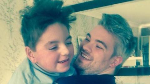 Paul Woodward his holding his son Kalel in his arms and they are both laughing. They are standing in front of a mirror in a living room.