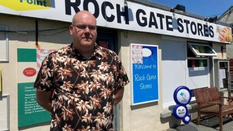 Tim, a bald white man wearing glasses and a Hawaiian shirt, standing outside the post office with a large sign on it saying 'Roch Gate Stores'