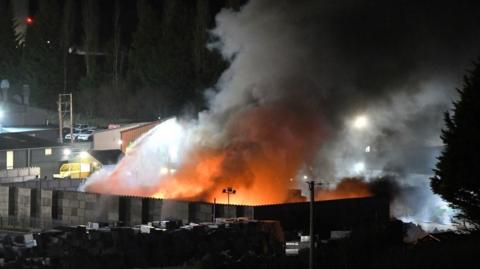 A large recycling plant with the main body of the building completely consumed by fire. Emergency services vehicles are visible to the side of the building.