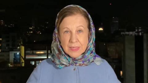 Lyse Doucet in a blue coat and headscarf, she is stood with a dark Tehran skyline behind her