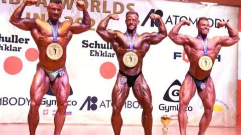 Three body builders pose with their arms raised wearing only trunks and medals around their necks.