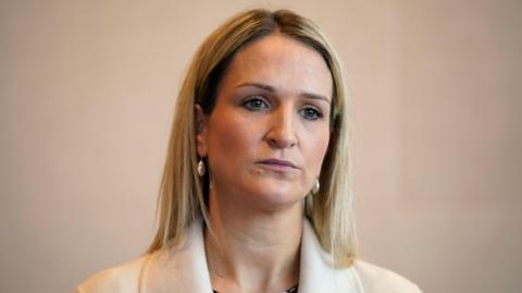 Helen McEntee - A woman with blonde, shoulder-length hair wears earrings and a white suit jacket. She is standing in front of a blank, neutral colour wall.