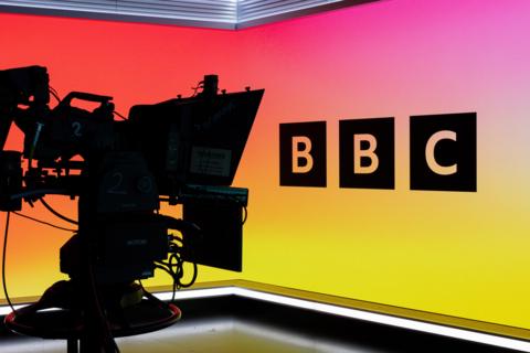 A stock image showing a broadcast studio camera in silhouette in front of a studio screen, it has a BBC logo on it and the colour scheme fades from red/pink at the top to yellow at the bottom