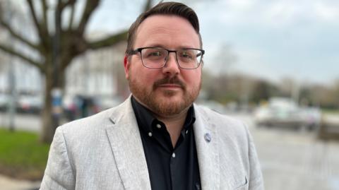 A man in a cream-coloured blazer and black shirt with glasses and beard looks at the camera.
